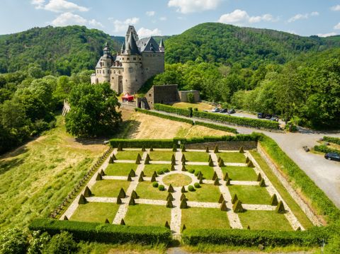 Eine beeindruckende Burg umgeben von grünen Hügeln. Im Vordergrund ist ein geometrischer Garten mit kunstvoll angelegten Hecken zu sehen.