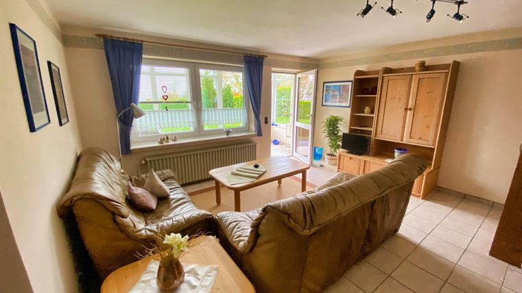 Ein gemütliches Wohnzimmer mit einem beigen Sofa und einem Holz-Couchtisch. Große Fenster lassen viel Tageslicht herein und bieten Ausblick auf den Garten.