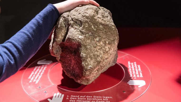 Ein großer, runder Stein liegt auf einem Tisch. Eine Hand berührt den Stein, während Anweisungen im Hintergrund sichtbar sind.