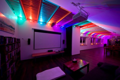 Ein moderner Raum mit farbigen LED-Lichtern an der Decke und einer Leinwand für Präsentationen. Der Boden ist aus Holz und es gibt bequeme Möbel.