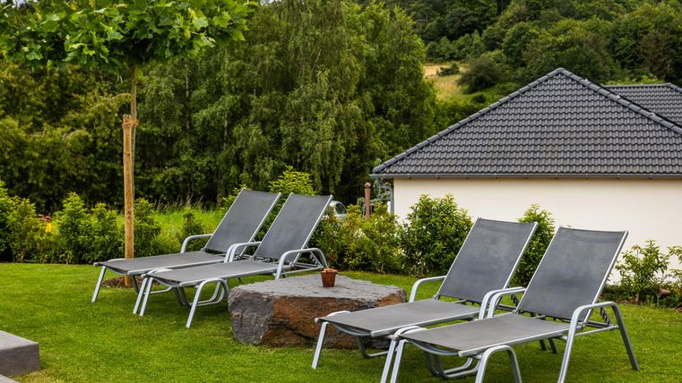Eine ruhige Terrasse mit mehreren Liegestühlen auf grüner Wiese. Im Hintergrund sind Bäume und ein Haus zu sehen.