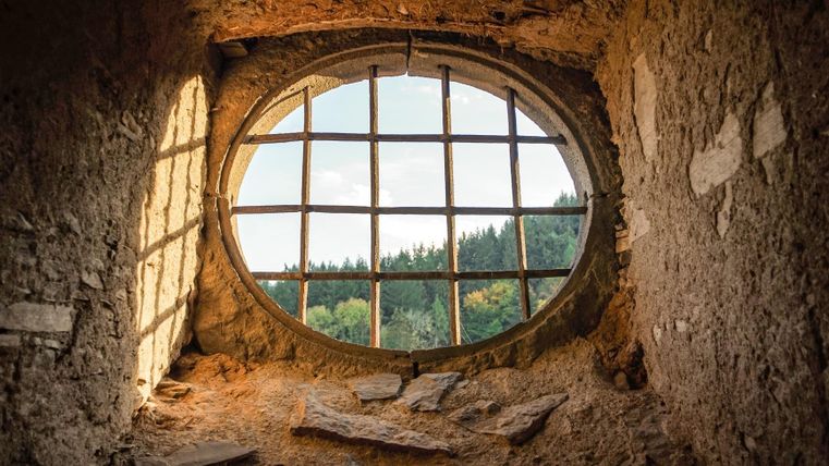 Ein rundes, vergittertes Fenster in einer alten Wand. Draußen ist eine grüne Landschaft mit Bäumen sichtbar.