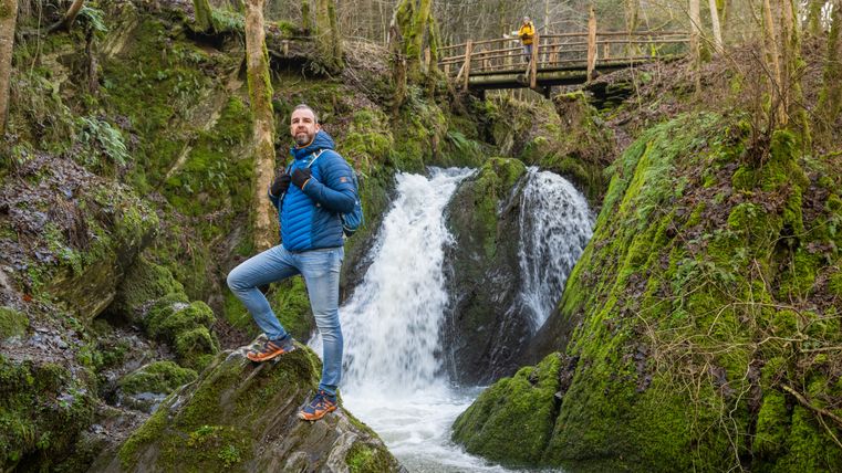 Wanderer mit Rucksack auf einem Felsen vor dem Wasserfall „Die Rausch“ im Tal der Wilden Endert, im Hintergrund eine Holzbrücke über der moosbewachsenen Schlucht.