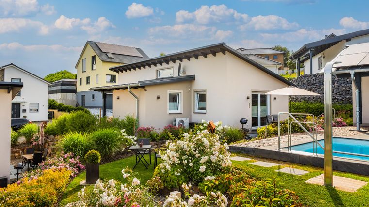Ein modernes Haus mit einem schönen Garten und einem Pool. Die Umgebung ist grün mit blühenden Pflanzen und blauem Himmel.