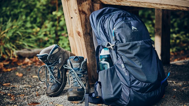 Ein Paar Wanderschuhe lehnt an einer hölzernen Bank, daneben ein blauer Rucksack mit einer Trinkflasche in der Seitentasche; herbstliches Laub liegt auf dem Kiesboden.