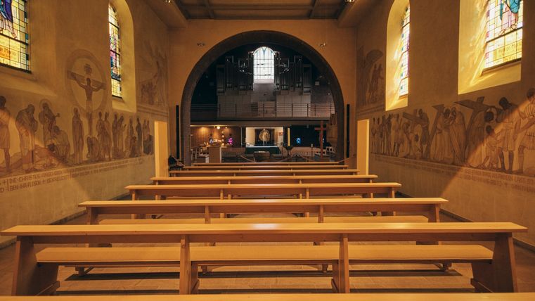 Blick durch das Kirchenschiff einer Kirche mit leeren Holzbankreihen. An den Wänden sind Kreuzweg-Darstellungen und bunte Kirchenfenster zu sehen. Im Hintergrund ist der Altarraum mit Orgel und Tabernakel sichtbar.