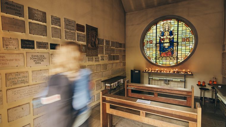 Kleine Kapelle mit einer Person in Bewegung im Vordergrund, umgeben von Dankestafeln an den Wänden. Im Zentrum ein rundes Buntglasfenster mit einer Marienfigur und ein Altar mit brennenden Kerzen