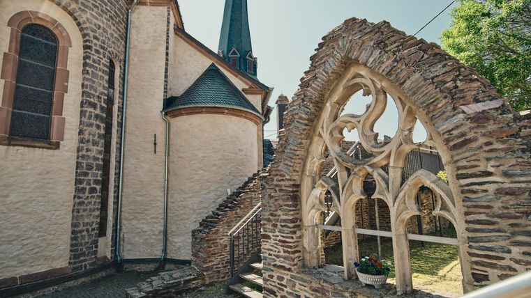 Eine alte Kirche mit einem spitzen Turm und gotischen Fenstern. Im Vordergrund ist ein historisches Mauerwerk zu sehen.
