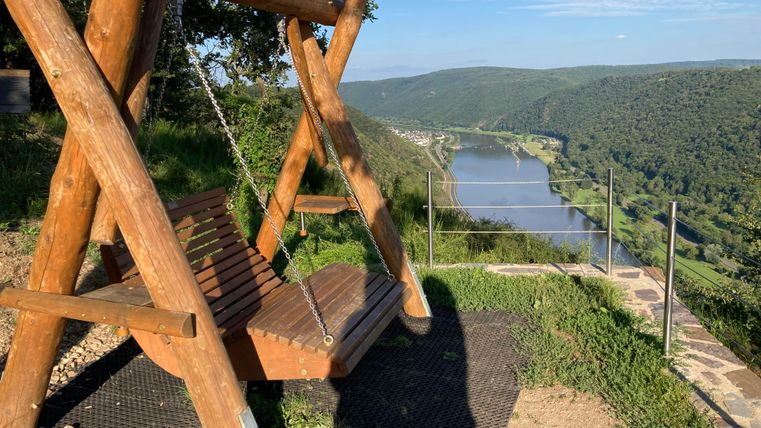 Eine Holzschaukel steht auf einem Hügel mit Blick auf einen Fluss. Die Landschaft ist grün und die Himmel ist klar.