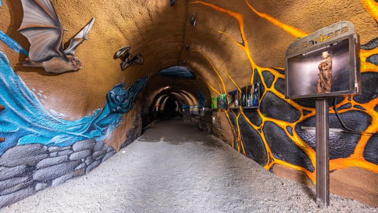 Ein kreativer Tunnel mit bunten Wandmalereien und informative Tafeln. Der Boden ist mit Kies bedeckt und die Beleuchtung sorgt für eine interessante Atmosphäre.