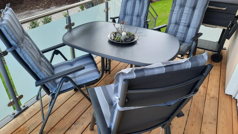 Ein Balkon mit einem schwarzen Tisch und vier passenden Stühlen. Auf dem Tisch steht eine kleine Pflanzenarrangement.