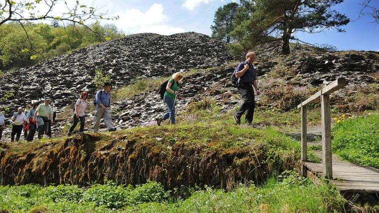 Eine Wandergruppe läuft über in Richtung einer kleinen Holzbrücke. Im Hintergrund sind Schieferhügel zu erkennen.
