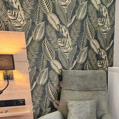 Ein gemütlicher Sessel vor einer Wand mit einem Pflanzenmuster. Das Ambiente ist warm und einladend.