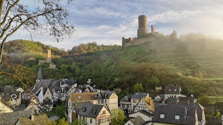 Eine malerische Landschaft mit einem historischen Dorf und einer Burg auf einem Hügel. Die Szene zeigt sanfte Hügel und eine ruhige Atmosphäre.