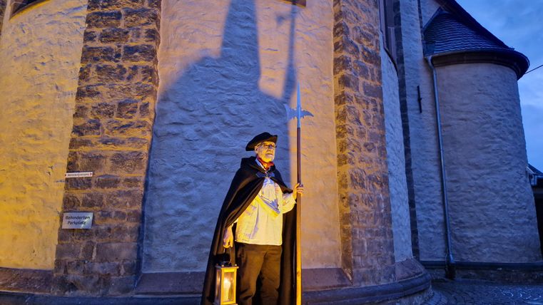Ein Mann in historischer Kleidung steht mit einer Laterne vor einer alten Kirche. Er trägt einen Umhang und hält einen Stock, während die Dämmerung einsetzt.