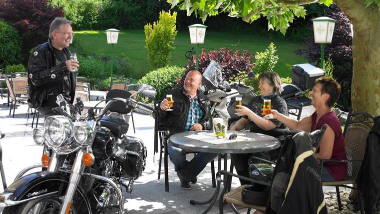 Eine Gruppe von Freunden sitzt an einem Tisch im Freien und genießt Getränke. Im Vordergrund steht ein Motorrad, und die Umgebung ist grün und einladend.