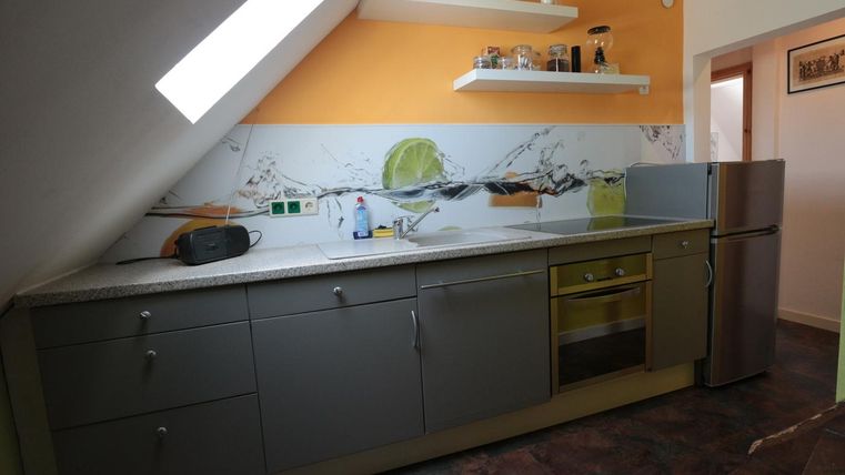 Eine Küche mit grauen Möbeln und einer bunten Wandgestaltung. Im Hintergrund befinden sich ein Kühlschrank und Regale mit verschiedenen Gegenständen.