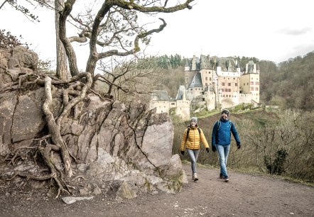 Zwei Wanderer in bunten Jacken gehen auf einem Pfad. Im Hintergrund ist die Burg Eltz zu sehen, umgeben von Wald., &copy; Eifel Tourismus GmbH, Dominik Ketz