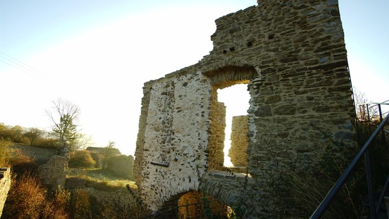 Ruinen einer alten Steinmauer umgeben von natürlicher Vegetation. Die Sonne scheint durch die Öffnungen und erzeugt eine malerische Atmosphäre.