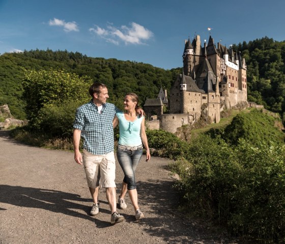 Ein Paar spaziert auf einem sonnigen Weg, im Hintergrund die malerische Burg Eltz, umgeben von gr&uuml;nen W&auml;ldern und blauem Himmel., &copy; Rheinland-Pfalz Tourismus GmbH, D. Ketz