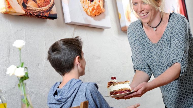 Eine lächelnde Frau serviert einem Jungen ein Stück Kuchen in einem gemütlichen Café. Die Wände sind mit einladenden Bildern von Gebäck dekoriert.