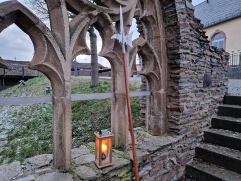 Ein historisches Tor aus Stein mit einem romantischen Lanternenlicht. Im Hintergrund sind grüne Wiesen und ein modernes Gebäude sichtbar.
