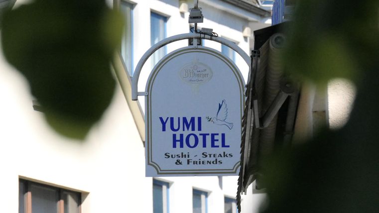 Ein Schild des Yumi Hotels, das Sushi, Snacks und Getränke anbietet. Es hängt an einem Gebäude in einer ruhigen Umgebung.