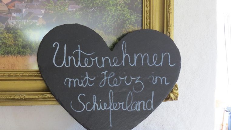 Ein herzförmiges Schild mit der Aufschrift "Unternehmen mit Herz in Schieferland". Das Schild ist auf einem Tisch neben einem gerahmten Bild platziert.