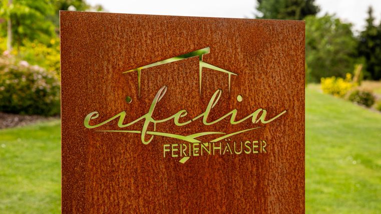Ein Schild mit der Aufschrift "eifelia Ferienhäuser" vor einer grünen Landschaft. Es ist ein einladender Ort für Urlaub und Entspannung.