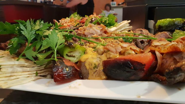 Ein üppiger Teller mit gegrilltem Fleisch, frischem Gemüse und Kräutern. Im Hintergrund sind Menschen in einem Restaurant zu sehen.