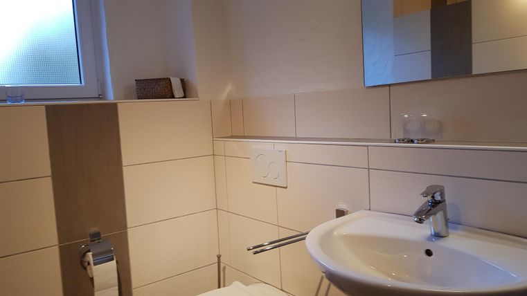 Ein modernes Badezimmer mit einer Toilette, einem Waschbecken und einem großen Spiegel. Die Wände sind hell gefliest und es gibt ein kleines Fenster für Tageslicht.