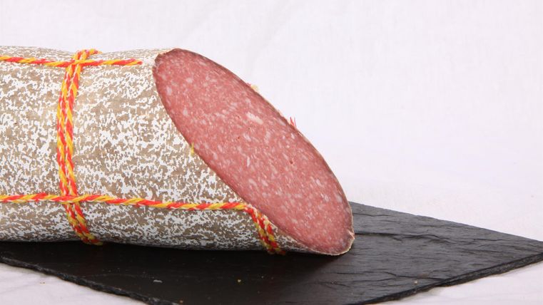 Eine lange Wurst mit einer grauen Oberfläche und roten Schnüren auf einem schwarzen Servierteller. Das Innere der Wurst ist rosa und frisch geschnitten.
