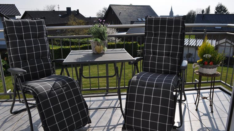 Eine gemütliche Terrasse mit zwei Stühlen und einem Tisch. Auf dem Tisch steht ein Blumentopf, und im Hintergrund sind grüne Wiesen und Häuser zu sehen.