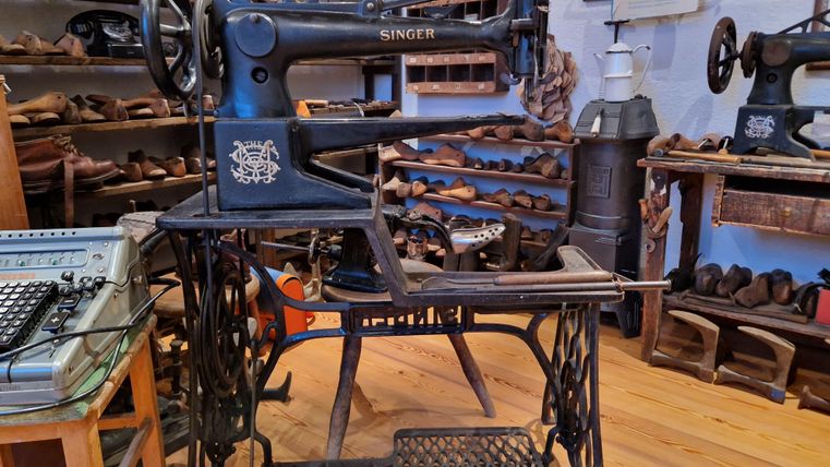 Eine alte Nähmaschine von Singer steht in einem Museum. Im Hintergrund sind viele Schuhe und andere Werkzeuge sichtbar.