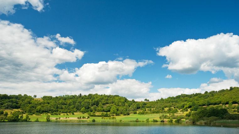 Ein ruhiger See umgeben von grünen Hügeln und Bäumen. Der Himmel ist blau mit einigen weißen Wolken.