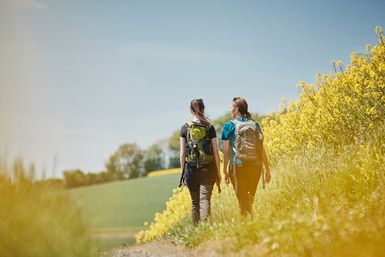 Zwei Frauen wandern entlang eines blühenden Rapsfeldes. 