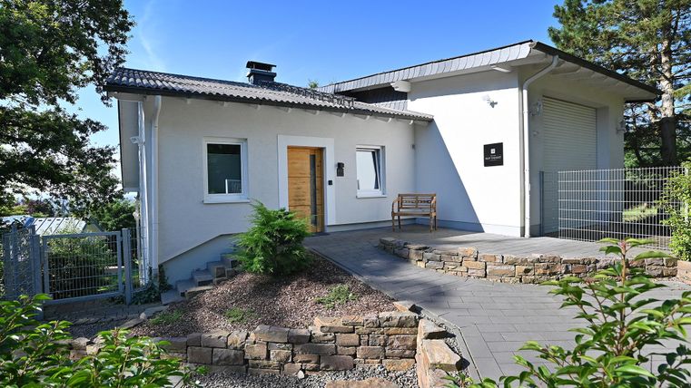 Ein modernes, helles Haus mit einem Garten und einer gepflasterten Auffahrt. Die Fassade ist weiß und es gibt Bäume in der Umgebung.