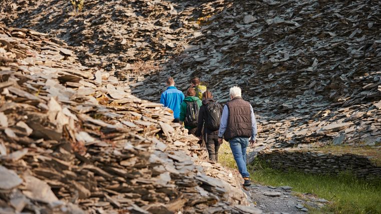 Eine Gruppe von fünf Personen wandert einen schmalen Pfad entlang, umgeben von steinigen Hängen. Die Landschaft ist grün und lädt zu einer Erkundungstour ein.