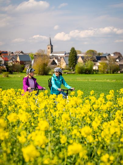 Two female cyclists are riding through a field of blooming rapeseed with a village in the background.