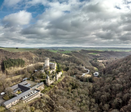 Luftaufnahme der Burg Pyrmont in der Eifel, umgeben von H&uuml;geln und W&auml;ldern. Der Himmel ist bew&ouml;lkt, die Landschaft weitl&auml;ufig und gr&uuml;n., &copy; Eifel Tourismus GmbH, D. Ketz