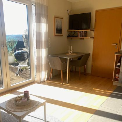 Ein gemütlicher Raum mit einem Tisch und zwei Stühlen. Große Fenster bringen viel Licht und bieten einen Blick auf den Außenbereich.