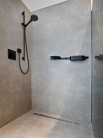 Eine moderne Dusche mit grauen Fliesen und schwarzer Armatur. Der Raum ist hell und minimalistisch gestaltet.