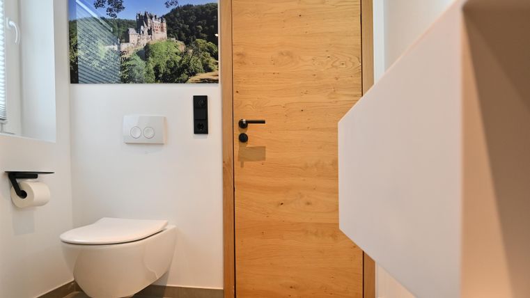 Ein modernes Badezimmer mit einer weißen Toilette und einer Holz-Tür. An der Wand hängt ein Bild einer Burg in einer grünen Landschaft.