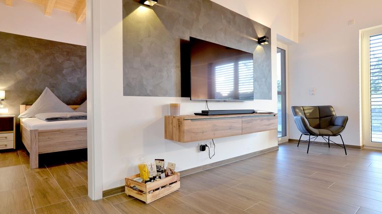 Ein modernes Wohnzimmer mit einem Fernseher an der Wand und gemütlichem Mobiliar. Die hellen Farben und Holzböden schaffen eine einladende Atmosphäre.