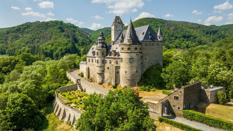 Eine beeindruckende Burg umgeben von grünen Wäldern und sanften Hügeln. Der Himmel ist blau mit einigen weißen Wolken.