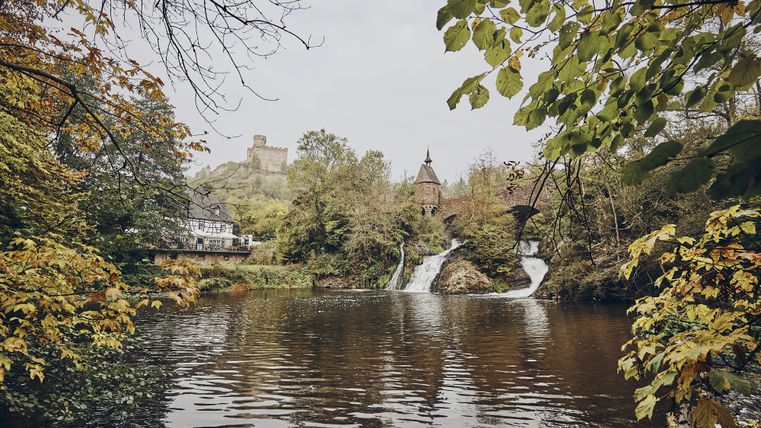Ein ruhiger Fluss mit einem Wasserfall und umgeben von bunten Herbstblättern. Im Hintergrund sind eine alte Burg und ein Haus sichtbar.