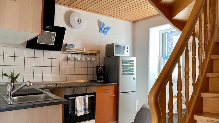 Eine moderne Küche mit Holzregalen und einer Treppe im Hintergrund. Die Ausstattung umfasst einen Herd, Kühlschrank und Küchenspüle.