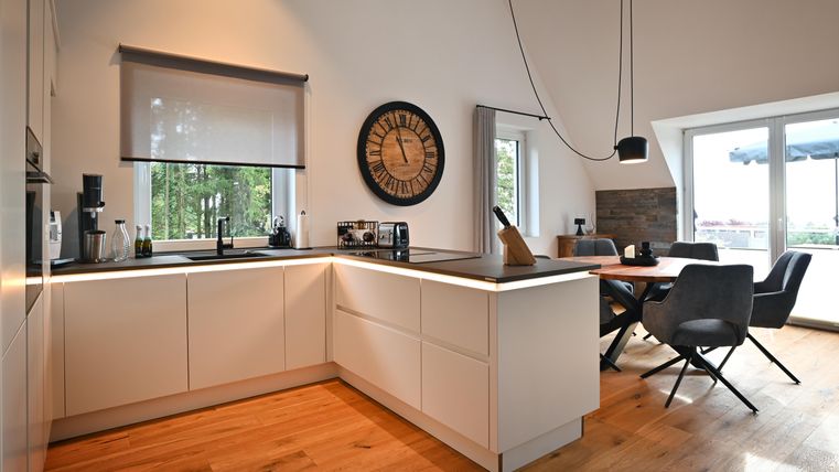 Eine moderne Küche mit einem hellen Holzfußboden und großen Fenstern. Am Tisch stehen komfortable Stühle, und eine Uhr ist an der Wand angebracht.