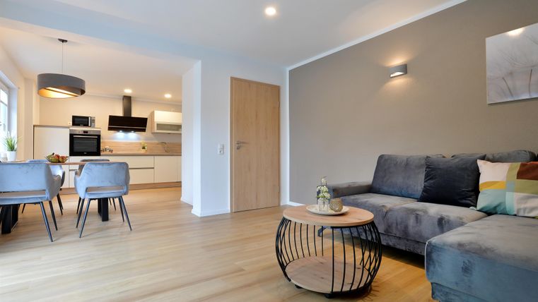 Ein modernes Wohnzimmer mit einem grauen Sofa und einem runden Tisch. Im Hintergrund ist eine offene Küche mit hellen Möbeln sichtbar.