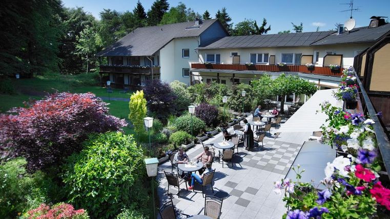 Ein schönes Hotel mit einem großzügigen Gartenbereich. Gäste sitzen draußen an Tischen unter Bäumen und genießen die sonnige Atmosphäre.